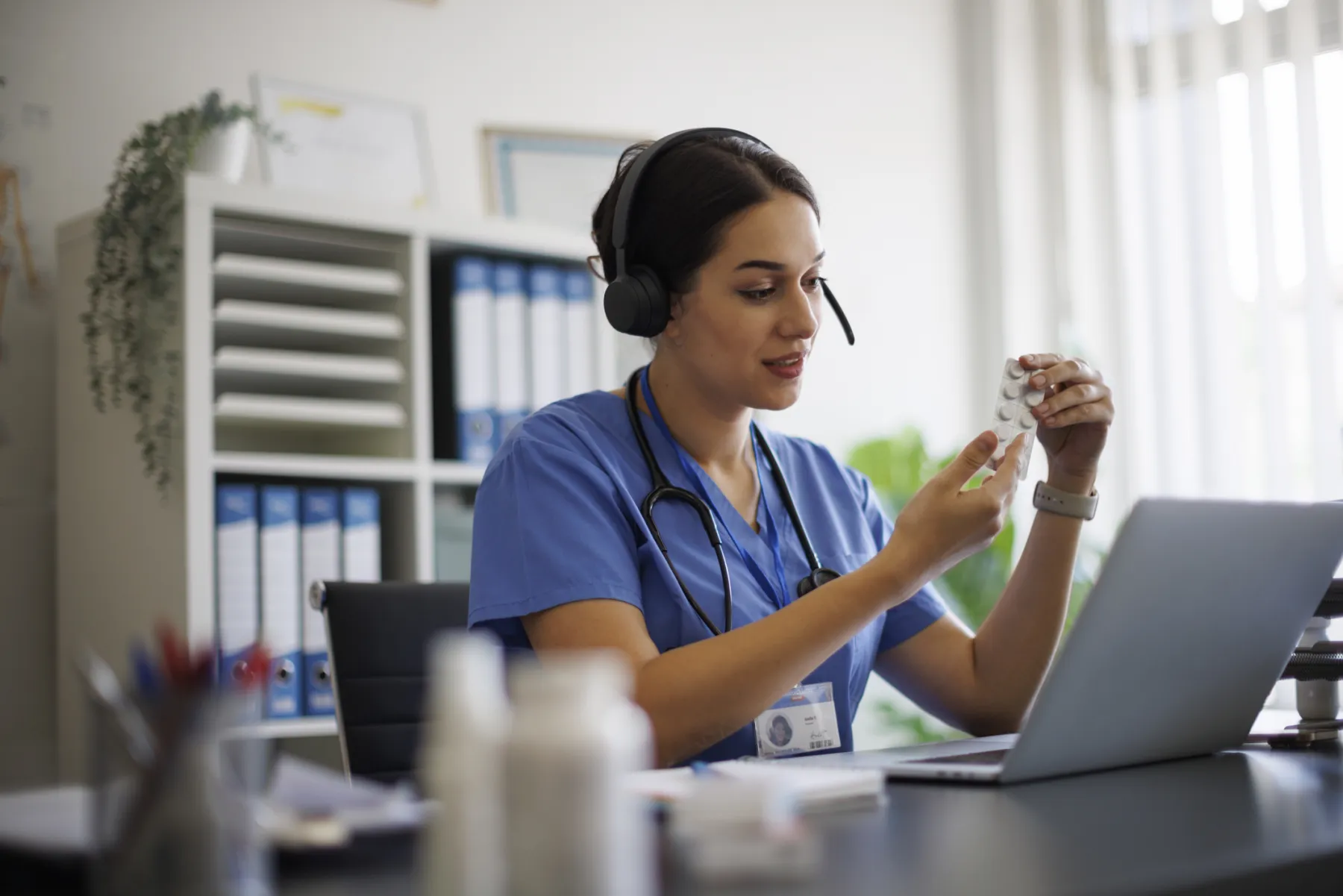 A healthcare agent in scrubs and a headset holding a medication pack at her laptop