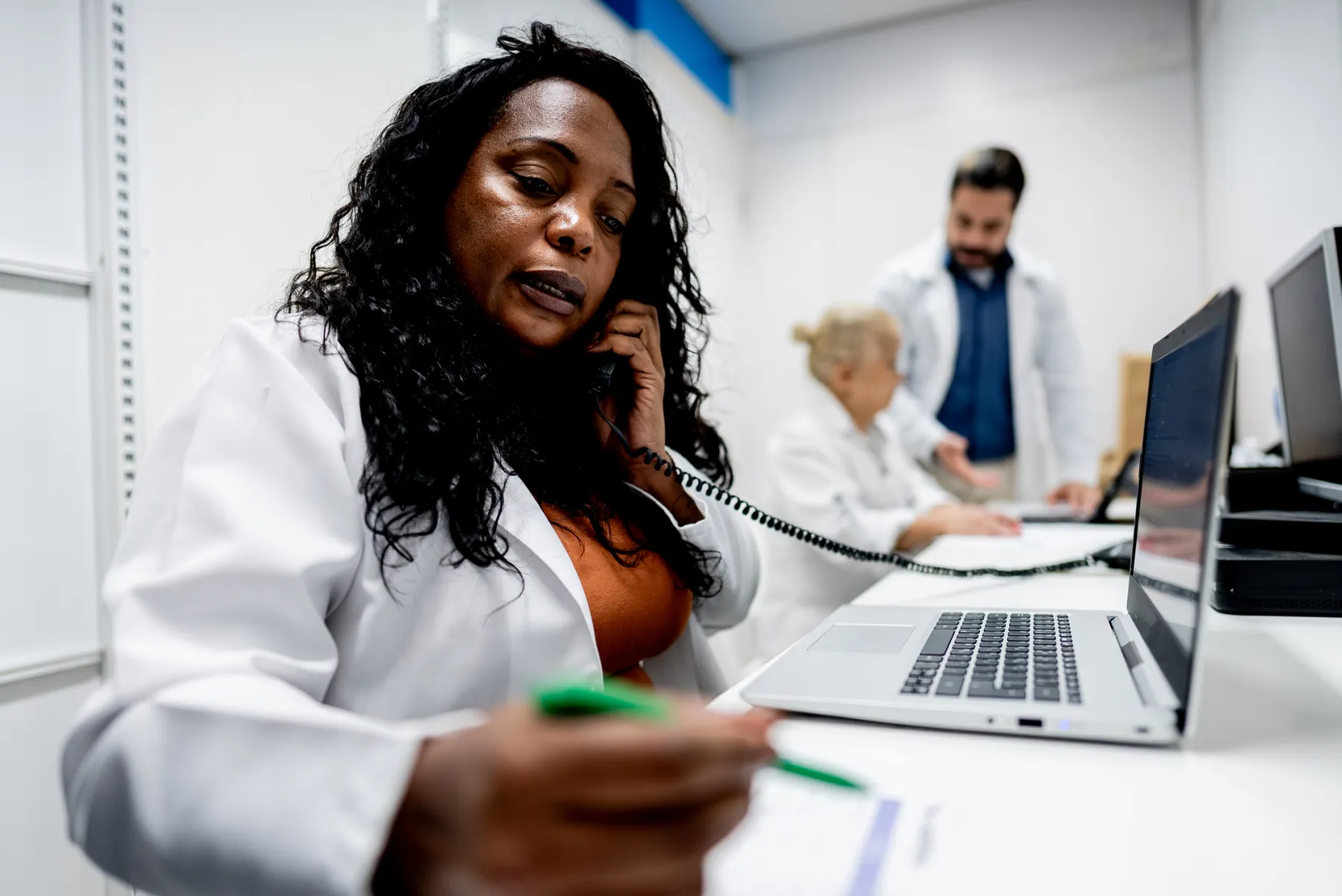A clinician in a white coat on the phone, taking notes at a laptop